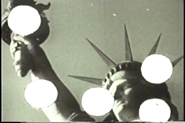 电影截图中的场景呈棕褐色，展示自由女神像的手臂和头部。五个白色圆点叠加于女神像上半身各处。