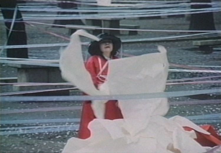 电影截图中的女子是艺术家草间弥生，她身穿一件飘逸的红色及膝长袍，头戴黑色宽边帽，周遭几株树干被多条丝带绕着。她手中拿着几片类似大白布的东西。