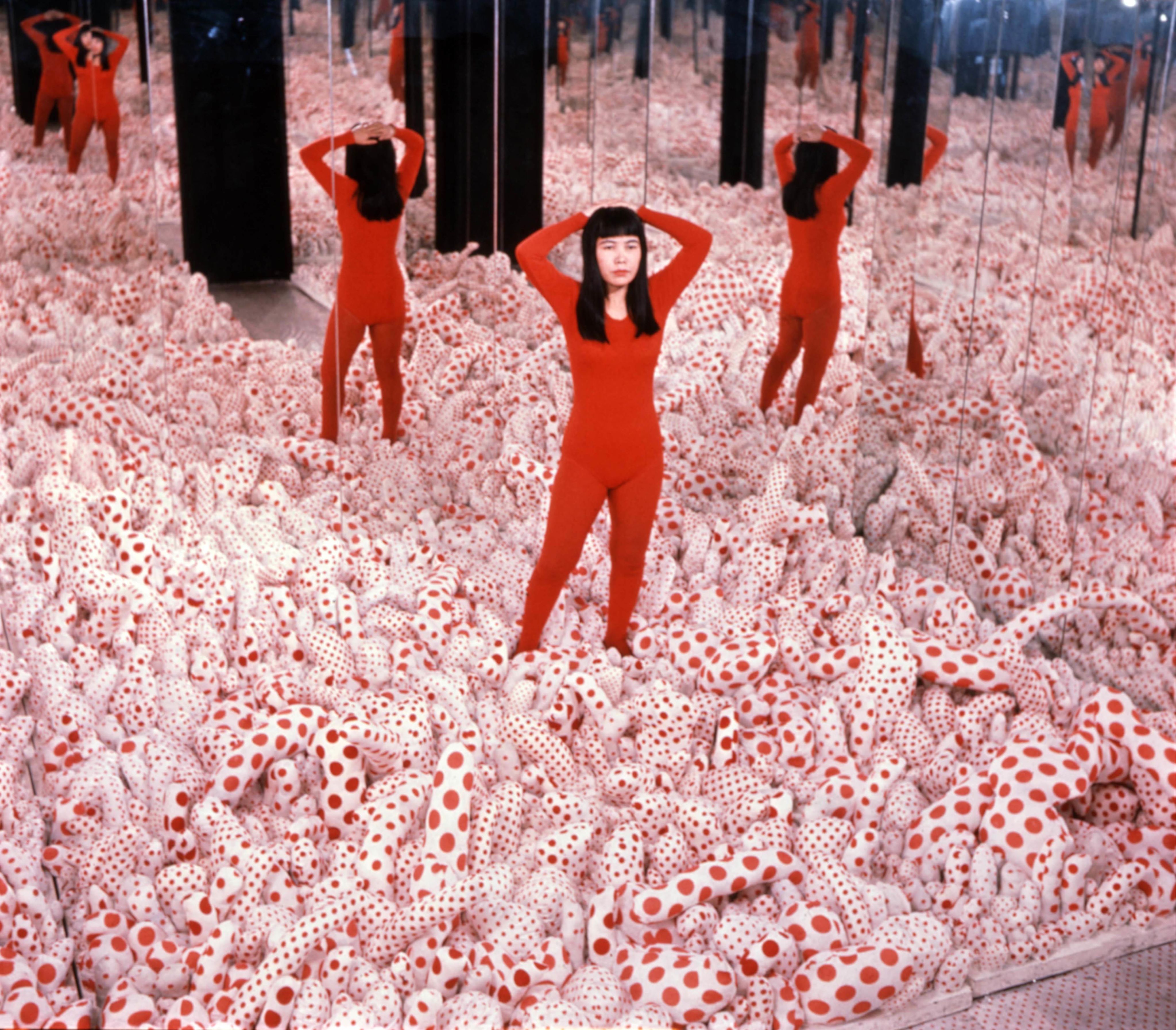 照片中，身穿红色贴身连衣裤的草间弥生站在室内，双手放在脑后。室内安装了数面墙壁般高度的镜子，地上满布带有红色圆点图案的突出状填充物。