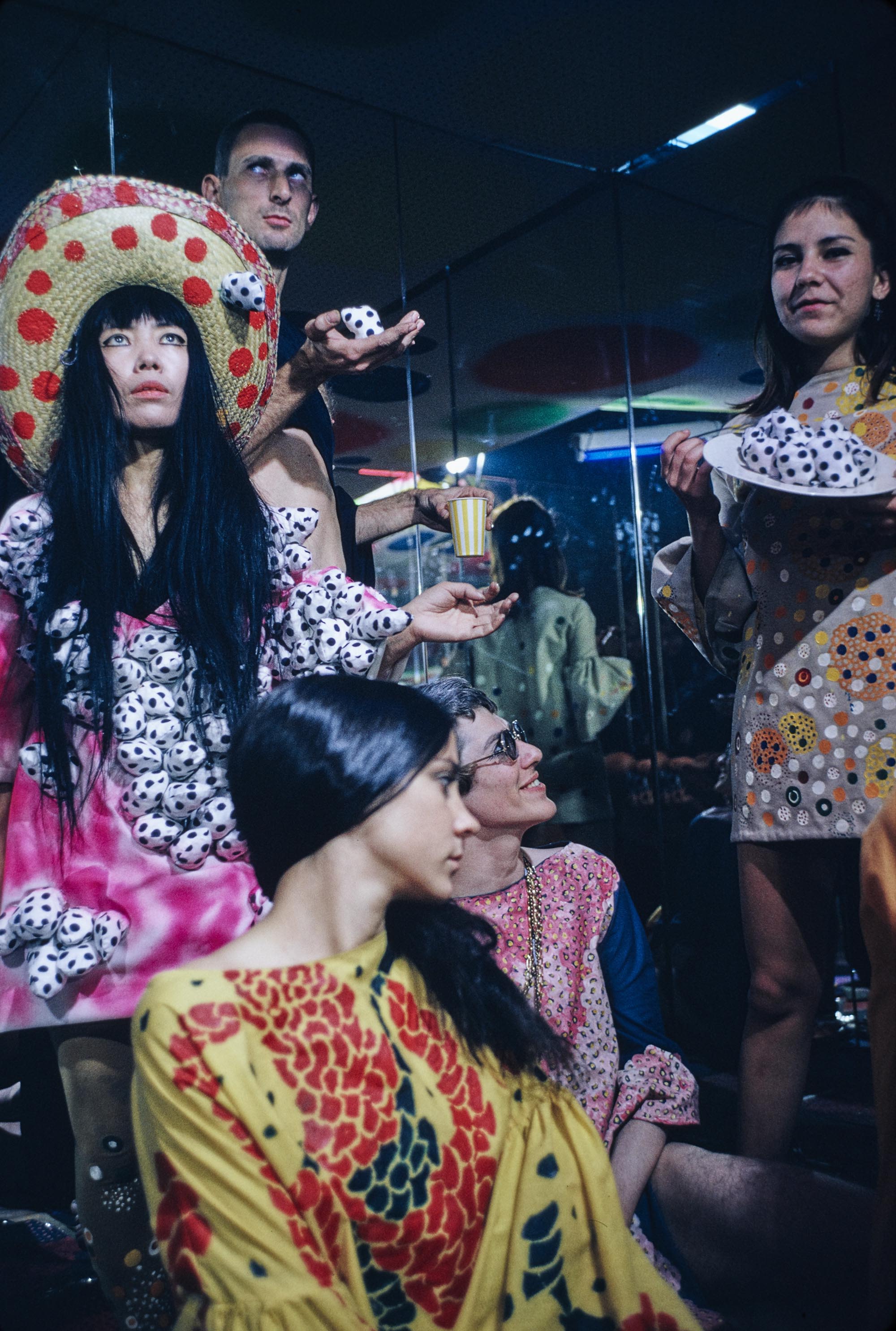 照片中，草间弥生和另外四人位于镜室内。草间头戴红色圆点草帽，身穿粉红色扎染连身裙，裙子表面被缀以黑色圆点的突出状填充物覆盖。前景里坐着两个人，其衣着打扮色彩缤纷。站在右边那人身穿点点图案连身裙，裙上缀以橙色和黄色圆形，另一个人则以手掌托着几件填充物软雕塑。