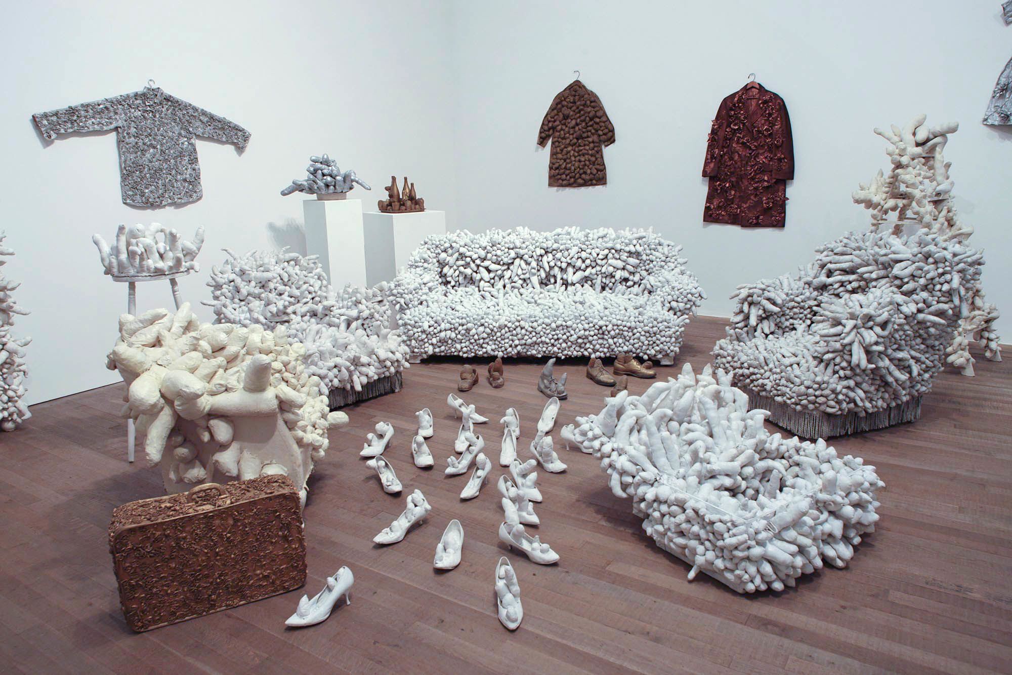 装置作品由几张沙发组成，每张沙发表面都有突出状填充物，并髹成白色或奶油色。墙上挂着几件大衣，前景的地上则放置了塞满突出状填充物的高跟鞋和一个行李箱。
