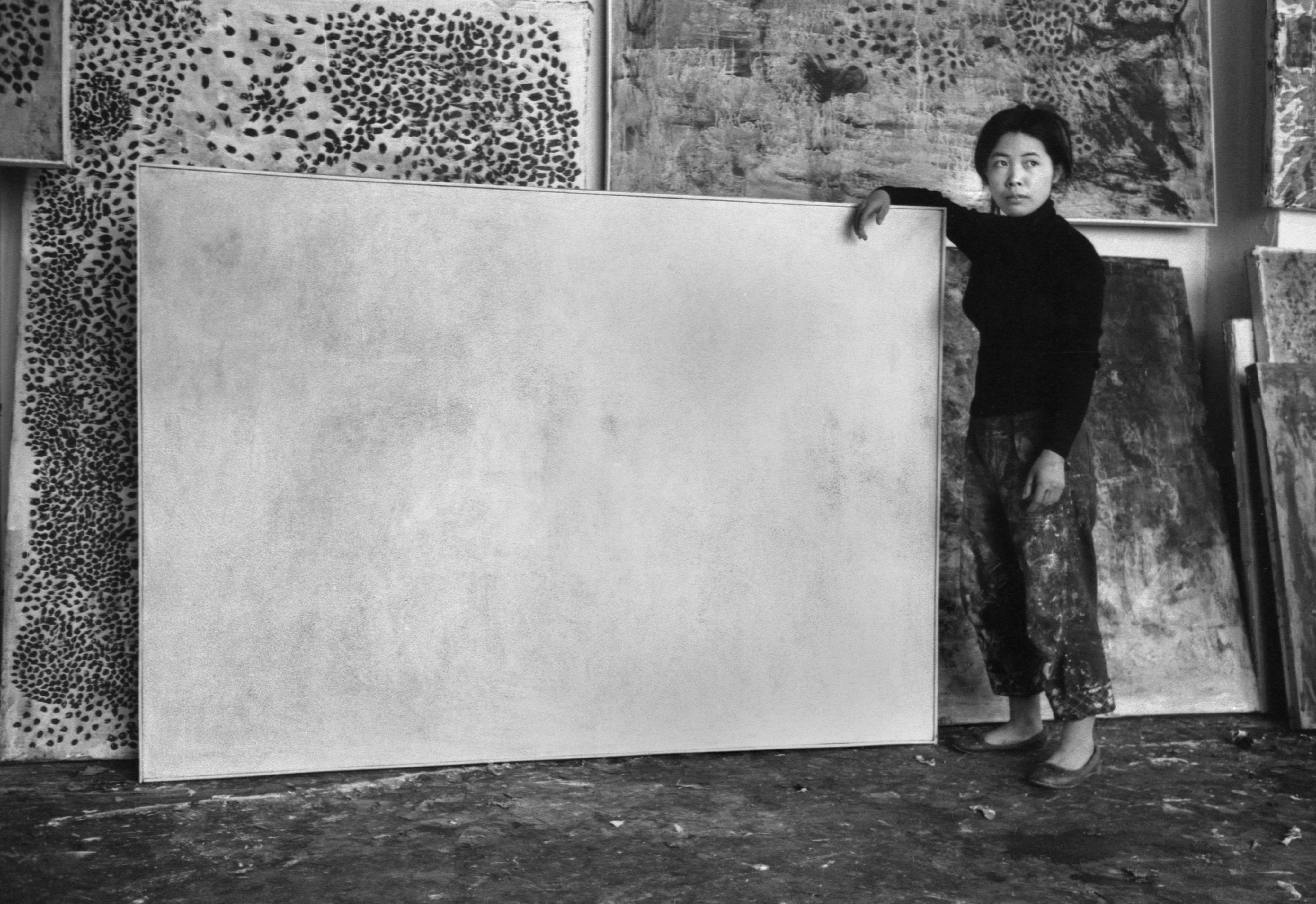 黑白照片中，草间弥生身处其纽约工作室内。她身穿黑色上衣和工装裤，凝视观者右方，并倚在一幅裱了框的横向作品上，画面呈现破格而抽象的着色效果。后方的墙上满是她绘上大量波点的作品，地上也搁着几幅画布。