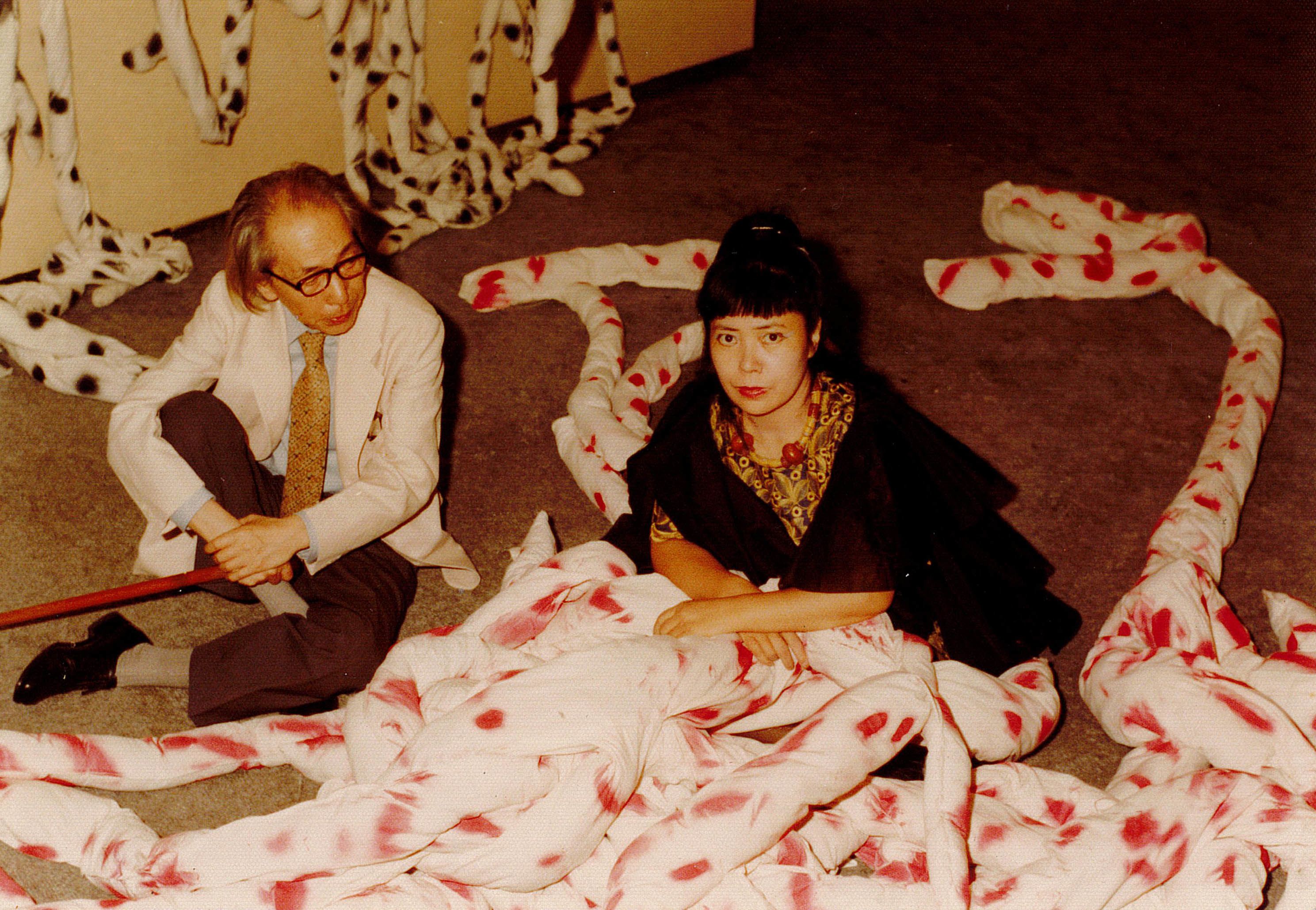 照片中，草间弥生与泷口修造一同坐在地板上，周围的红色圆点填充织物松散地缠绕在一起，形成一堆堆的状态。草间戴着一条粗大的串珠项链，望向镜头，泷口则穿着正式西服，手持拐杖，把目光投向她。